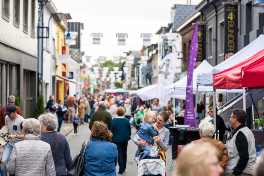Drukke straatmarkt met mensen, kraampjes en een moeder die een lachend kind vasthoudt. (vergroot foto)